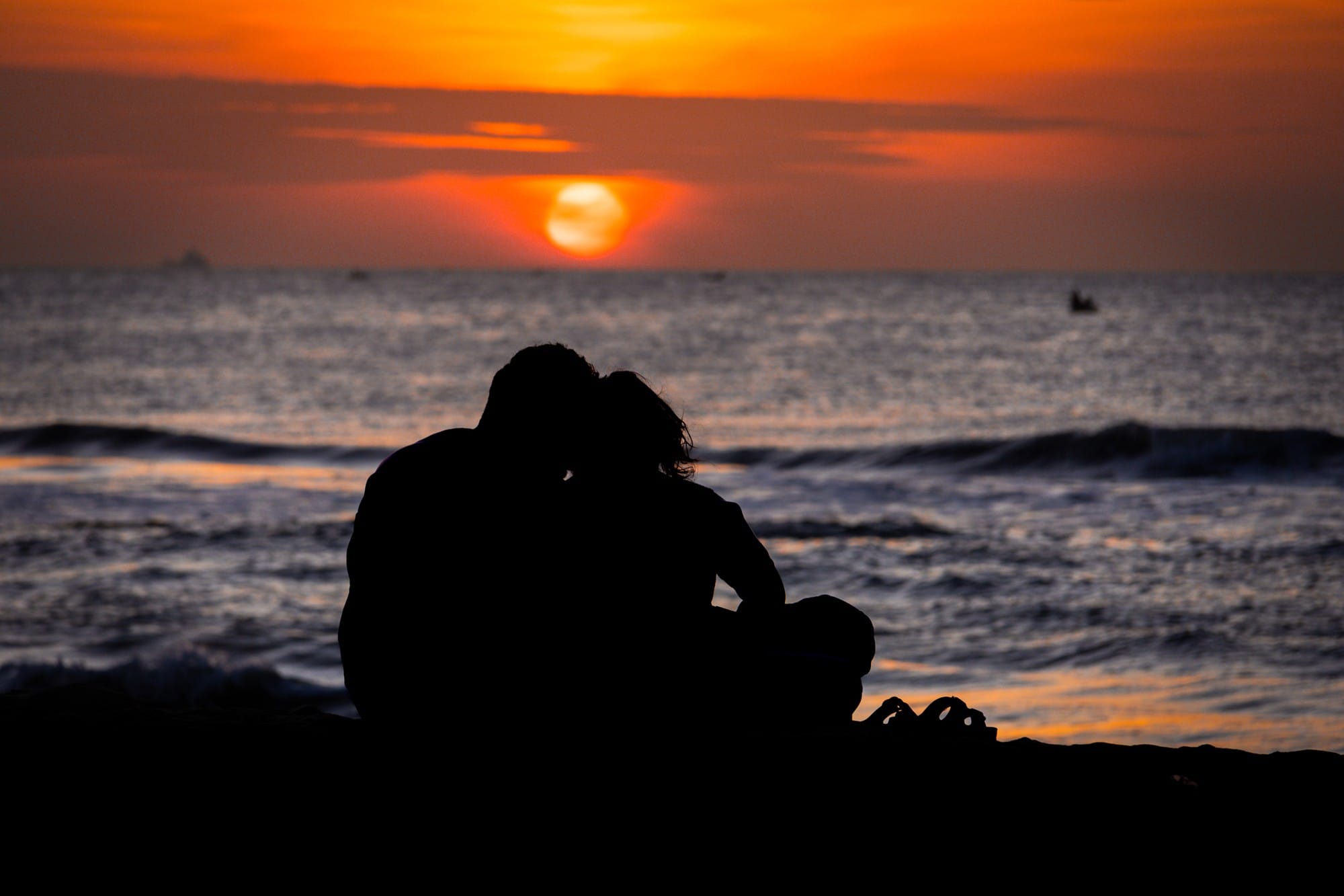 Silhouettes of two people sitting together beside the sea at sunset.