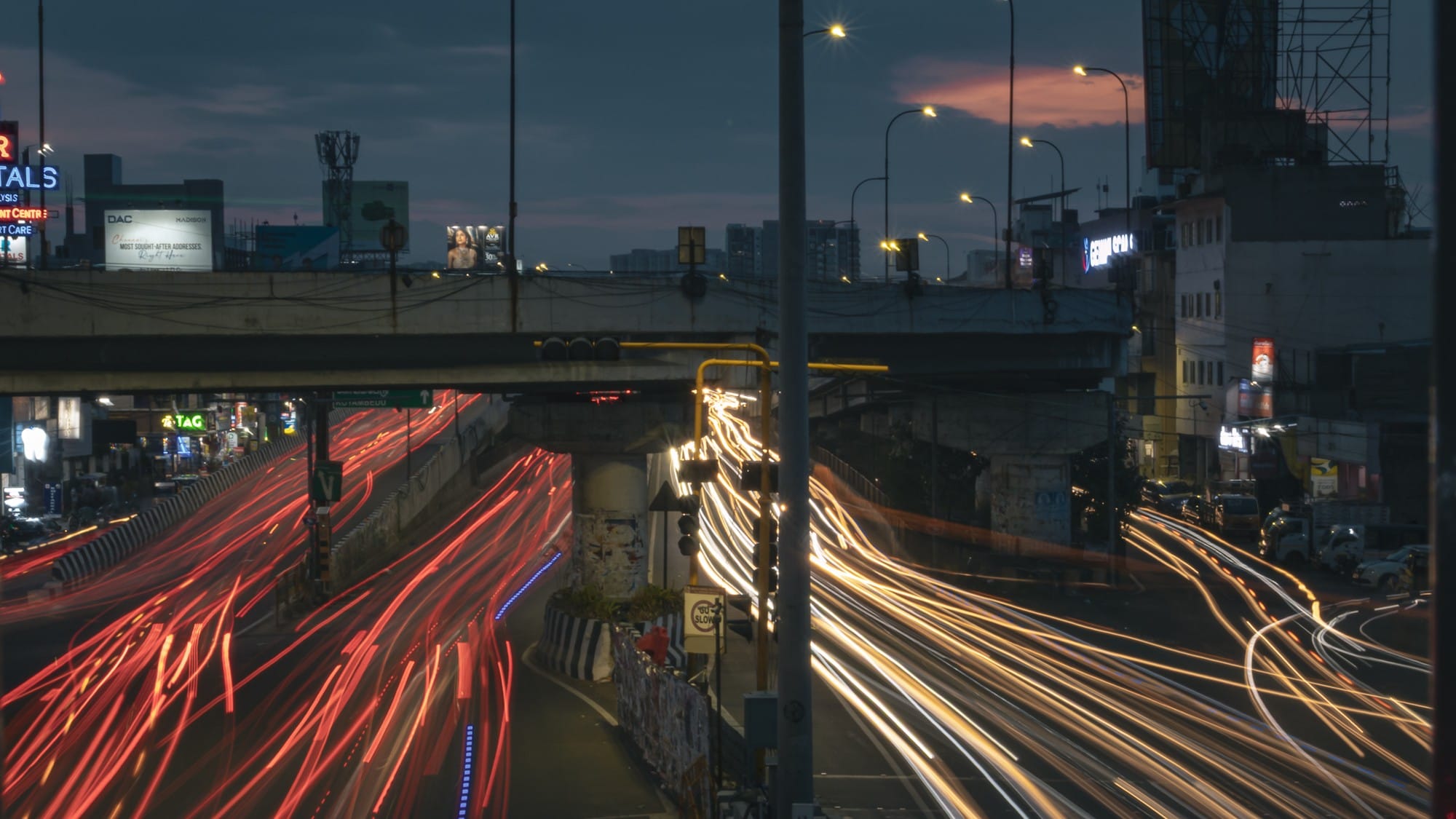 Long-exposure photograph of red and white traffic trails beneath a city overpass at dusk.