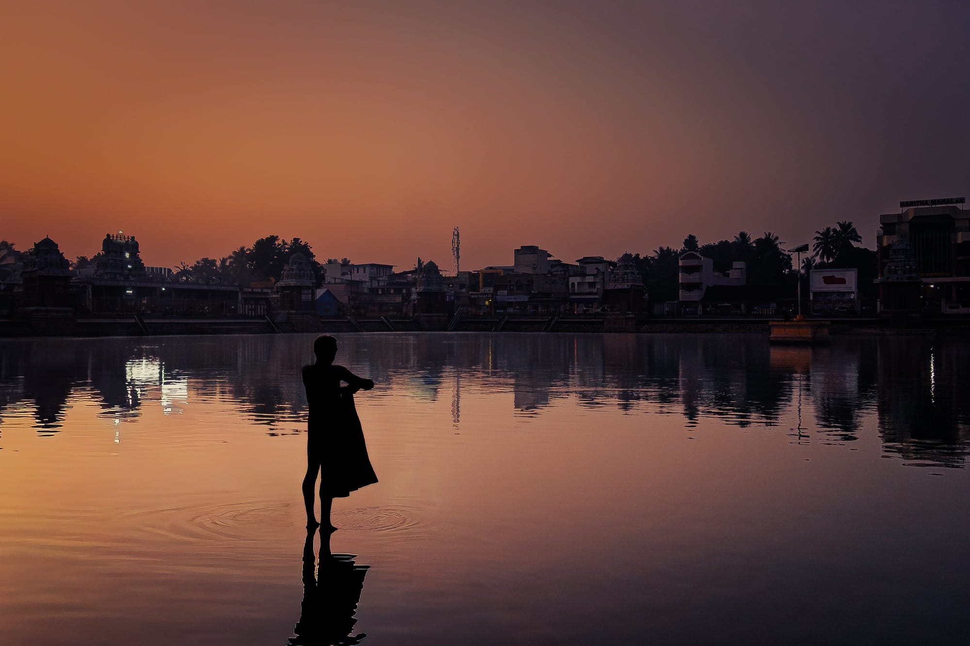 Silhouette of a person standing beside still water reflecting an orange evening sky.