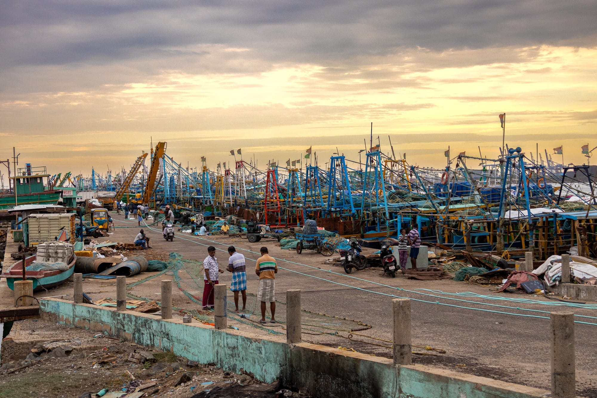 Busy fishing harbour and people on a quay beneath evening clouds.