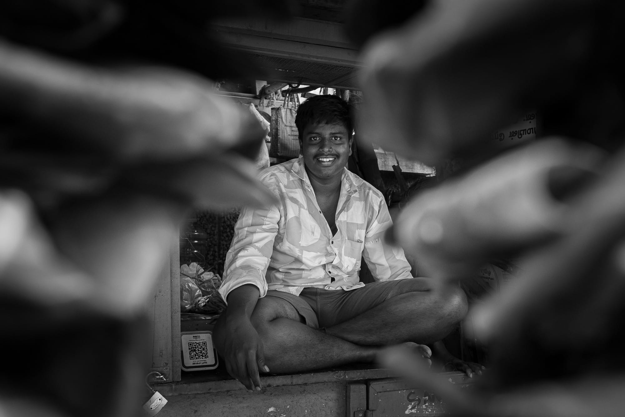 Smiling man at a market, framed by objects in the foreground in black and white.