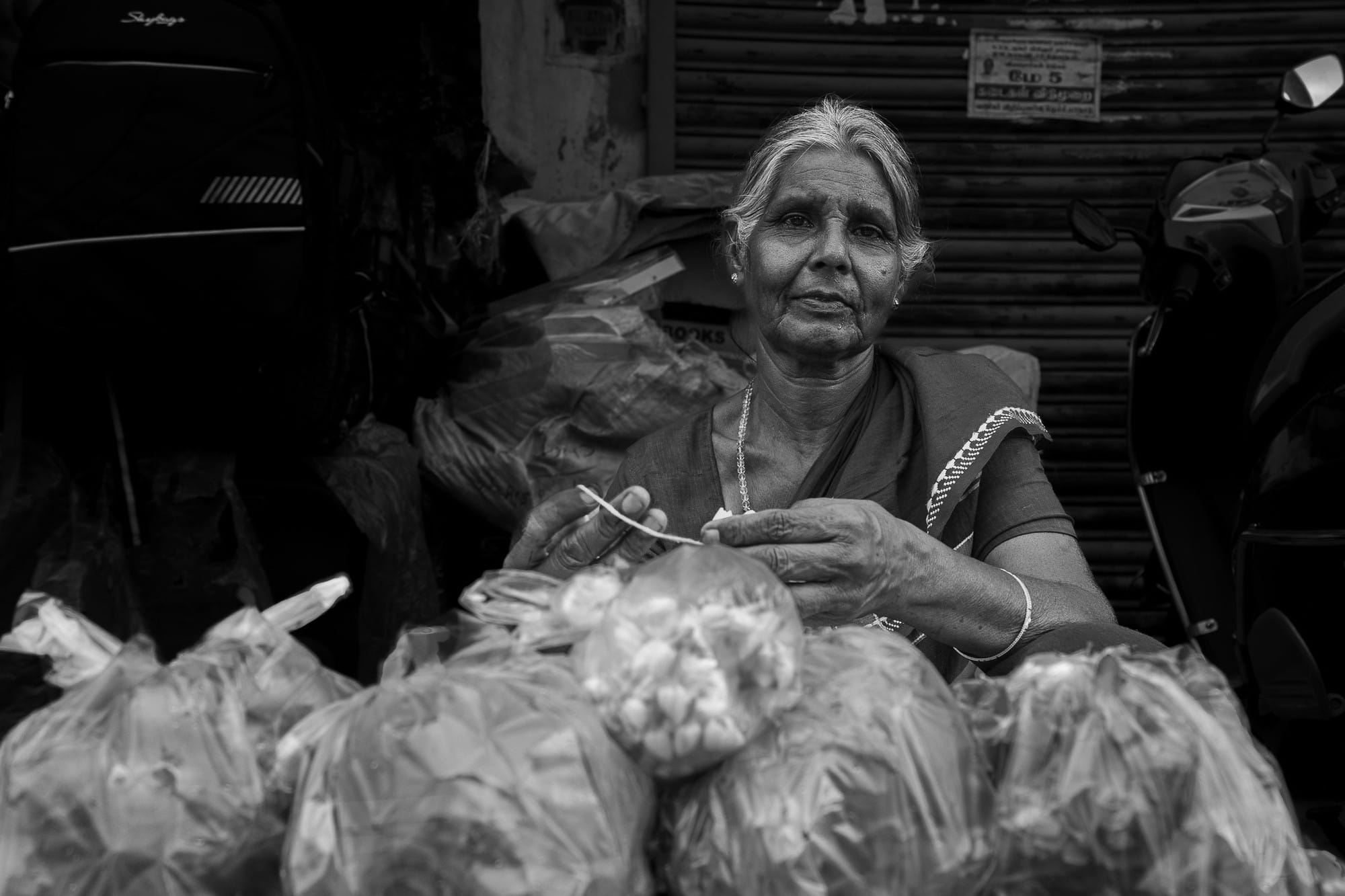 Black-and-white portrait of an older woman seated beside bags of market produce.