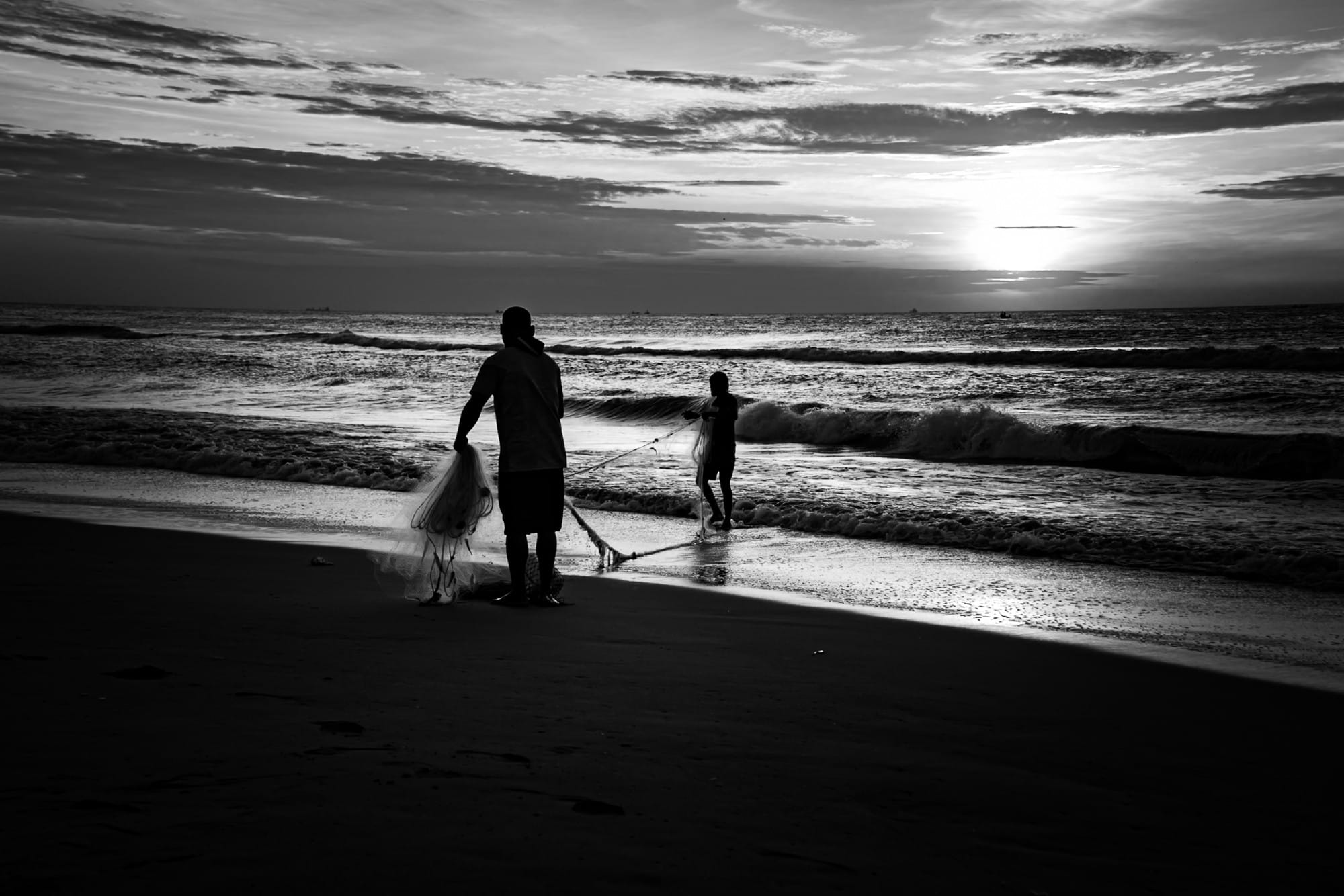 Black-and-white photograph of people walking with fishing nets at the shoreline.