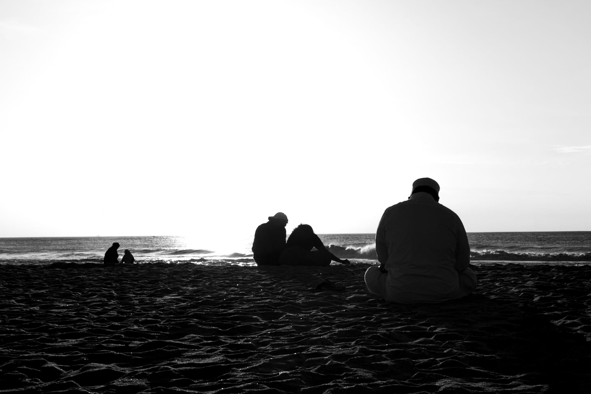 Black-and-white photograph of people sitting on the beach facing the bright horizon.